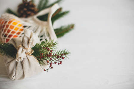 Zero waste christmas holidays. Stylish christmas gift wrapped in linen fabric with natural green branch and red berries on background of reusable cotton bag with oranges on white  table.の写真素材