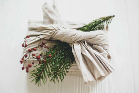 Stylish christmas gift wrapped in linen fabric and decorated with natural fir branch and red berries on white rustic table background. Zero waste christmas holidays.の写真素材