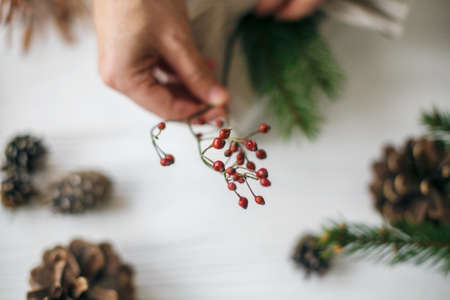 Red berries close up in hand, florist decorating stylish christmas gift in linen fabric with red berries branch on white rustic table with pine cones and green fir.の写真素材