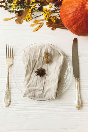 Eco friendly Thanksgiving feast. Stylish plate with cutlery, linen napkin, anise and autumn leaves, pumpkin,  autumnal flowers on white table. Rustic table setting decorの写真素材