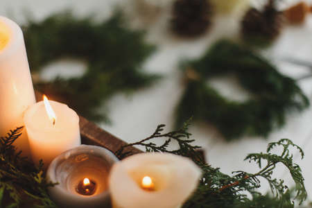 Burning candles on rustic background with christmas wreath, pine cones and ornaments on wooden table in evening. Holiday workshop advent, cozy atmosphere. Copy spaceの写真素材