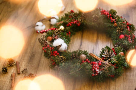 Christmas wreath on wooden rustic background in lights. Traditional christmas wreath with red berries and ornaments, pine cones, cotton and golden lights on wood, holiday decorの写真素材