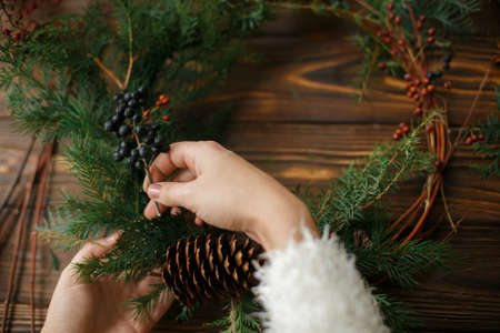 Making rustic christmas wreath. Hands holding berries and decorating natural cedar branches in wreath on rustic wooden background. Seasonal holiday workshop or making wreath at homeの写真素材
