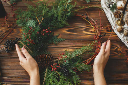 Modern rustic christmas wreath on wooden table, holiday advent. Female hands holding stylish christmas wreath with red berries, green cedar branches and pine cone. Making festive decorationの写真素材