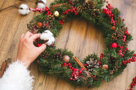 Person making traditional christmas wreath on rustic wooden background. Florist hand decorating christmas wreath with cotton, red berries, ornaments and pine cones. Holiday workshopの写真素材