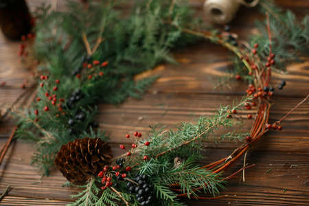 Rustic christmas wreath on wooden table, holiday advent. Modern christmas wreath with red berries, green cedar branches and pine cone. Making stylish festive decoration at workshopの写真素材