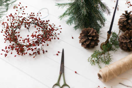 Making simple rustic christmas wreath on white wooden table at home, holiday advent. Christmas wreath with red berries, thread, scissors, pine cone and candle on rustic backgroundの写真素材