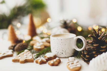 Christmas gingerbread cookies, coffee in stylish white cup, pine cones  and warm lights on white wooden table. Hello winter, cozy moody image with selective focusの写真素材