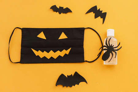 Evil face mask, disinfection gel bottle, bat and spider decorations on orange background. Halloween 2020 safe celebration flat lay. Stay creative Stay safe due coronavirus pandemicの写真素材