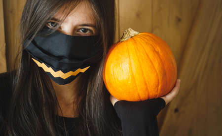 Halloween 2020, celebrating in safe way with social distance during coronavirus pandemic. Young woman in black face mask with jack o lantern smile holding pumpkin on wooden backgroundの写真素材