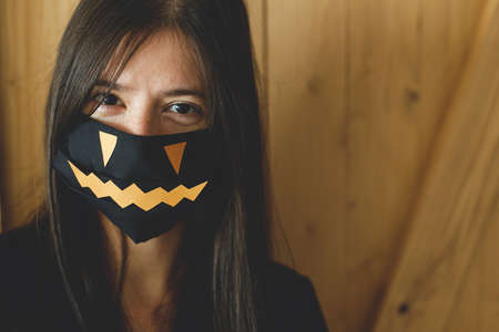 Halloween 2020 celebration with safety measures and social distance due coronavirus pandemic. Young woman in black face mask with jack o lantern smile on wooden background. Stay safeの写真素材