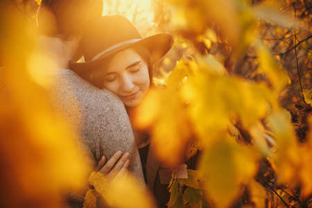 Stylish sensual couple gently hugging among golden autumn leaves in warm sunset light. Young fashionable woman hugging man close up, embracing at autumn tree. Romantic momentの写真素材