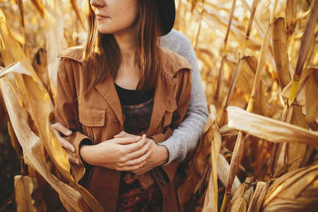 Young man gently hugging his fashionable woman, touch close up among golden corn leaves. Happy stylish couple embracing in autumn corn field in warm sunset light. Romantic sensual momentの写真素材