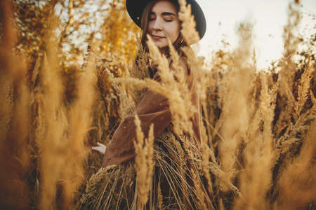 Stylish boho woman posing among grass in autumn field in warm sunset. Young fashionable female in brown coat and hat relaxing in herbs. Carefree authentic momentの写真素材