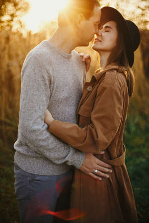 Happy stylish couple embracing in autumn meadow in warm sunset light. Romantic sensual moment. Young fashionable woman and man kissing in autumn fieldの写真素材
