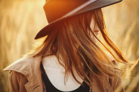 Carefree boho woman with windy hair in sunset light in autumn field. Young stylish female in hat relaxing on background of grass and herbs in sunshine. Sensual happy momentの写真素材