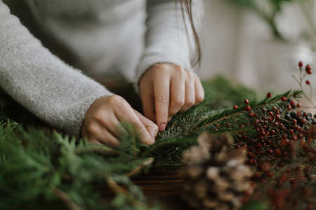 Close up of female hands holding green fir branches and making rustic christmas wreath on background of rustic wooden table with pine cones, berries. Handmade festive decor, holiday adventの写真素材