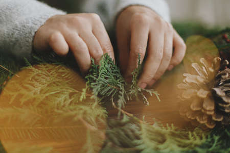 Close up of female hands holding cedar branches and making rustic christmas wreath on background of rustic wooden table with pine cones, berries and festive lights. Handmade festive decorの写真素材