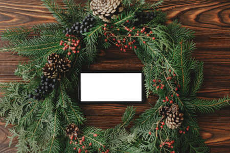 Phone with empty blank screen in rustic christmas wreath on wooden table flat lay. Festive app,...