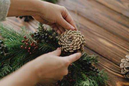 Making rustic christmas wreath, seasonal winter workshop. Florist hands holding pine cone and making christmas wreath on wooden table with natural festive decorations. Holiday adventの写真素材