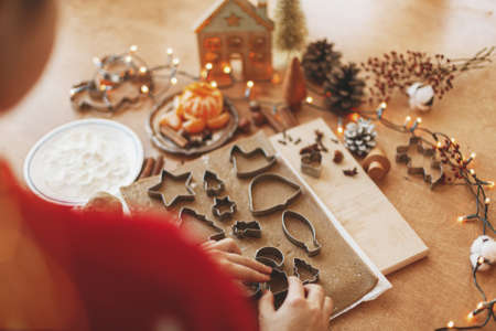 Hands cutting gingerbread dough with festive metal cutters on rustic table with spices, oranges,...