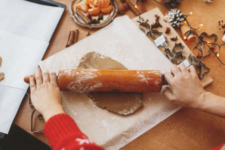 Hands rolling raw gingerbread dough with wooden rolling pin on background of metal cutters, festive decorations and lights on rustic table. Person making gingerbread cookies, Christmas adventの写真素材