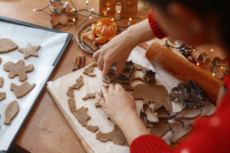 Hands cutting gingerbread dough with festive metal cutters on rustic table with spices, oranges,...