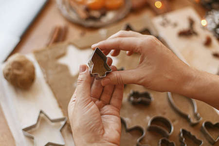 Hands cutting gingerbread dough with christmas tree metal cutter on rustic table with spices,...
