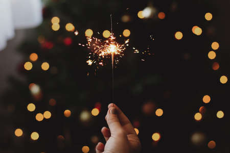 Happy New Year! Burning sparkler in female hand on background of christmas tree with lights in dark...