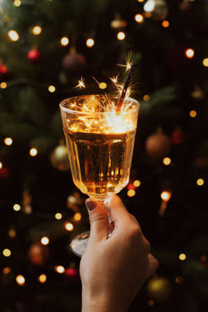 Happy New Year! Burning firework in champagne glass in hand on background of christmas tree with lights in dark festive room. Celebrating new year, stylish aesthetic image.の写真素材