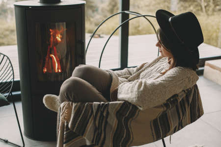Stylish woman in knitted sweater and hat warming up at modern black fireplace with view on mountains. Cozy warm moments at cold season. Hipster female relaxing in comfortable homeの写真素材