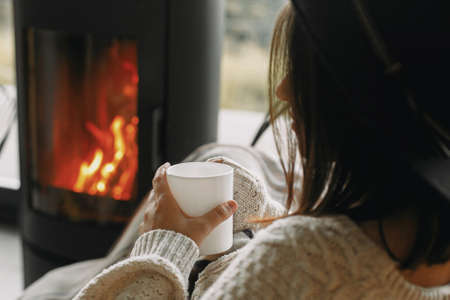 Stylish woman in knitted sweater relaxing with warm cup of tea at modern black fireplace with view...