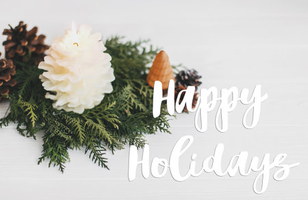 Happy holidays text handwritten on christmas pine cone candle and tree in rustic christmas wreath on...
