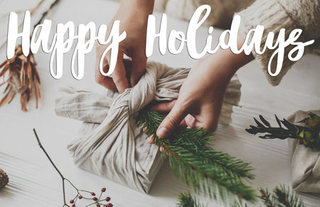 Happy holidays text handwritten on hands wrapping stylish furoshiki christmas gift in linen fabric on rustic table with fir and pine cones, zero waste. Greeting card, modern seasons greetingsの写真素材