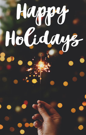 Happy holidays text handwritten on burning sparkler in hand on background of golden bokeh lights in festive dark room. Greeting card, modern seasons greetingsの写真素材