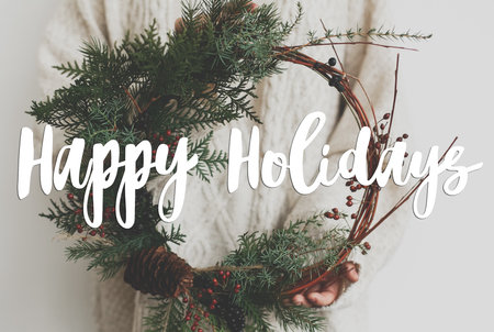 Happy holidays text handwritten on woman in sweater holding modern christmas wreath on white background. Greeting card, modern seasons greetingsの写真素材