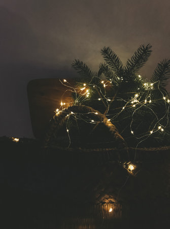 Stylish wicker basket with Christmas tree branches in illumination lights on rustic wooden chair in...