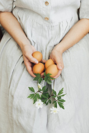 Happy Easter! Easter eggs and spring flowers in hands of woman in rustic linen dress. Woman holding natural easter eggs and spring blooms. Aesthetic eco holiday, stylish imageの写真素材