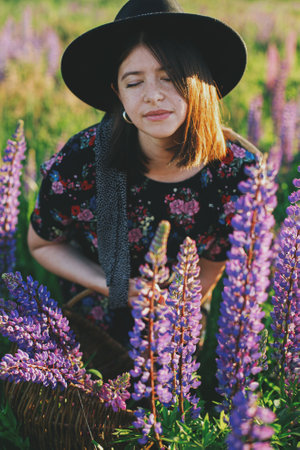 Beautiful woman relaxing in sunny lupine field, holding rustic basket with flowers. Tranquil...