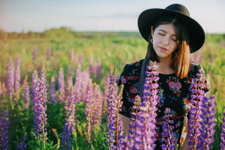 Beautiful stylish woman relaxing in sunny lupine field and holding rustic basket with flowers. Tranquil atmospheric moment. Young female in vintage dress and hat gathering wildflowers in countrysideの写真素材
