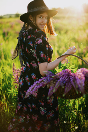 Happy beautiful woman walking in sunny lupine field, holding rustic basket with flowers. Tranquil atmospheric moment. Young female relaxing in countryside meadow at sunset.の写真素材