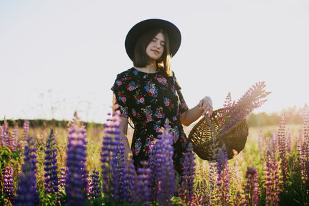 Beautiful stylish woman relaxing in sunny lupine field and holding rustic basket with flowers. Tranquil atmospheric moment. Young female in vintage dress and hat gathering wildflowers in countrysideの写真素材