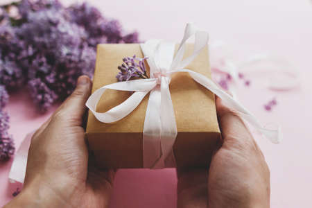 Hands holding gift box with ribbon and lilac flowers on pink paper. Happy mothers day and valentine's day congratulations concept. Purple lilac flowers bouquet with craft present box.の写真素材