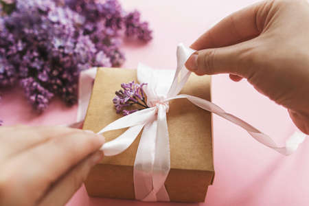 Happy mothers day and valentines day concept. Hands wrapping gift box with ribbon and lilac flowers on pink paper. Purple lilac flowers bouquet with craft present boxの写真素材