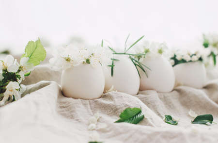 Natural eggs in cute floral wreaths on linen fabric background with blooming spring branch and white...