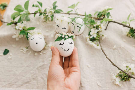 Easter egg with drawn cute face in floral wreath in person hand on background of natural eggs and...