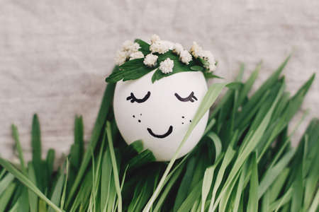 Easter egg with drawn sleeping face in floral wreath in grass on rustic linen fabric background....