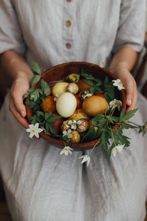 Woman in linen dress holding easter eggs and spring flowers in wooden bowl. Stylish easter and quail eggs in natural dye and spring blooms. Aesthetic holiday. Happy Easter!の写真素材