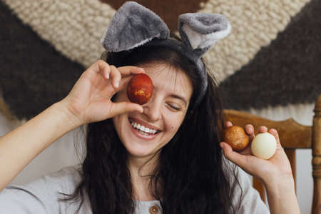Happy Easter! Happy young woman in bunny ears and linen dress holding easter eggs at eyes and...