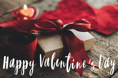 Happy Valentine's day text on stylish gift box with red ribbon and red velvet hearts on rustic wood. Love concept. Valentines day greeting cardの写真素材
